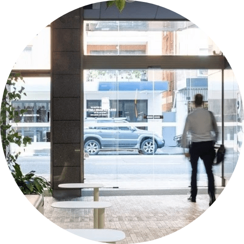 A man is exiting an office building through a glass door. the glass is immaculate, tables are cleaned. Office cleaning services include cleaning of glass window and common areas like in this picture of office space with plants and tables